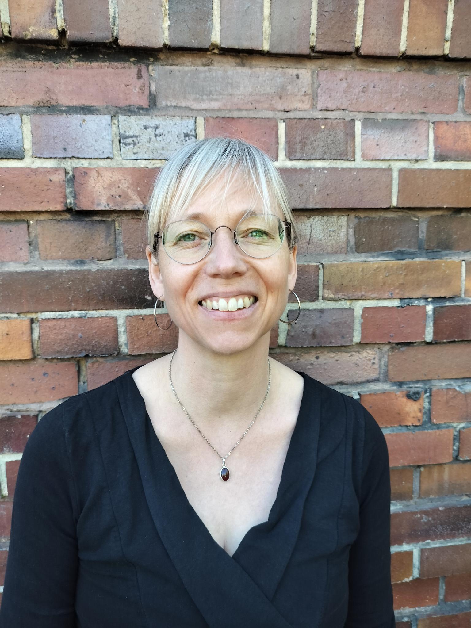 Maxie steht vor einer Backsteinwand. SIe trägt ein schwarzes Oberteil, große Ohrringe und eine kleine Kette. Ihre blonden Haare sind zum Zopf gebunden. Maxie trägt eine dünne Brille und lächelt in die Kamera.