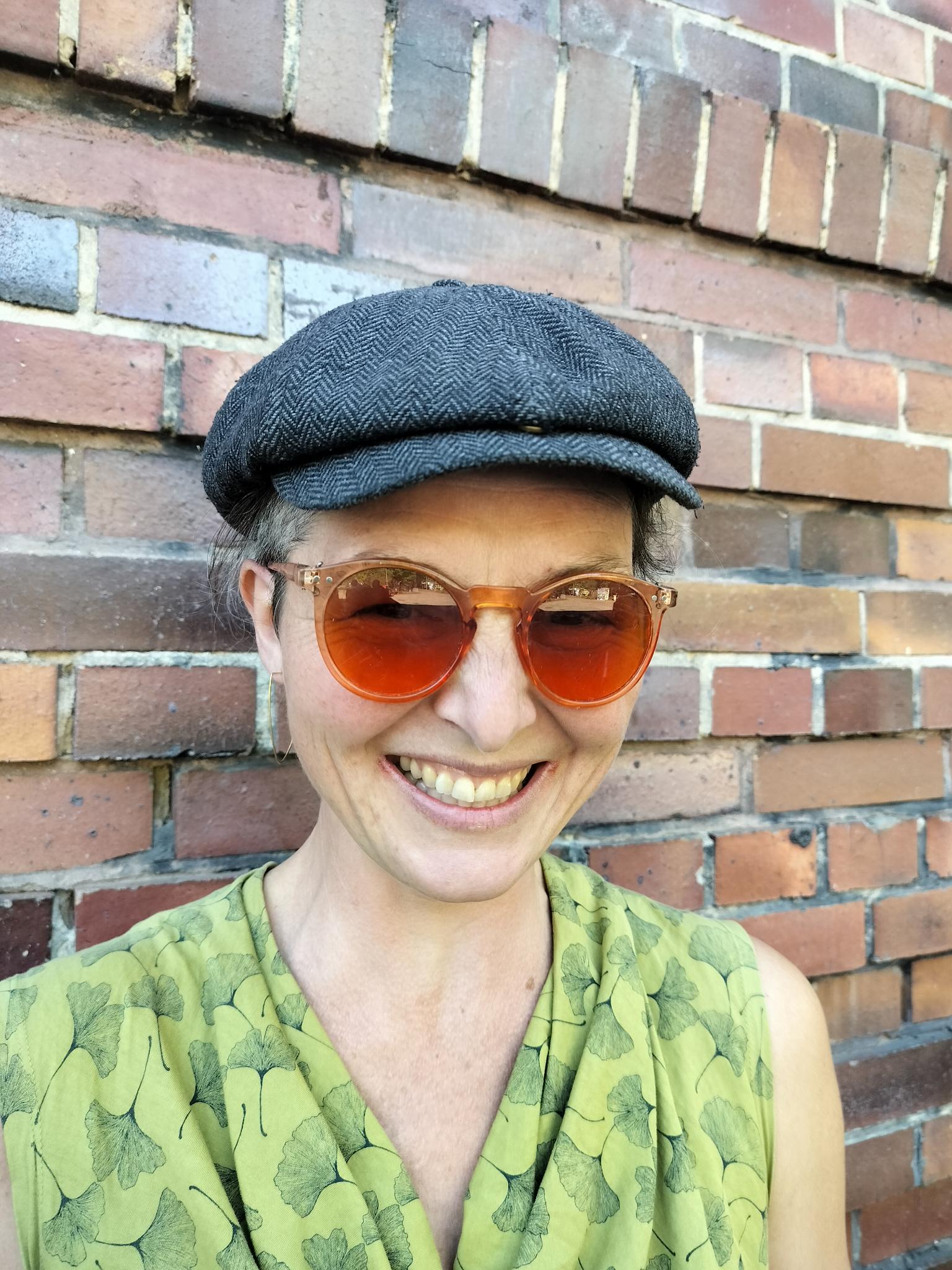 Jolika trägt eine graue Kappe, eine orangefarbene Sonnenbrille und ein grünes Oberteil. Sie steht vor einer Backsteinwand und lacht in die Kamera.