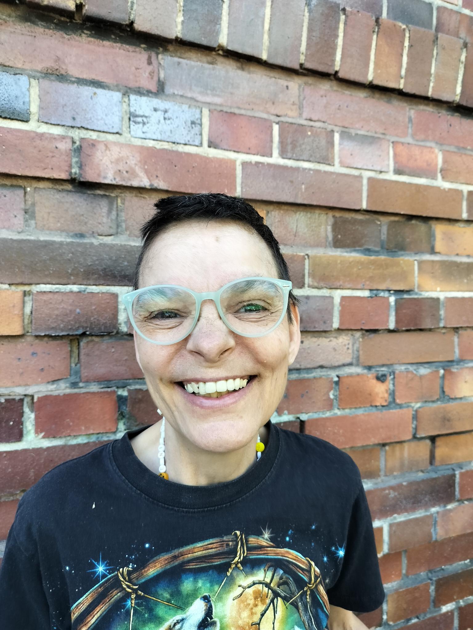 Christine trägt kurze dunkelbraune Haare, ein schwarzes T-Shirt mit Aufdruck und eine hellgrüne Brille. Sie lacht in die Kamera. Hinter ihr ist eine Backsteinwand.