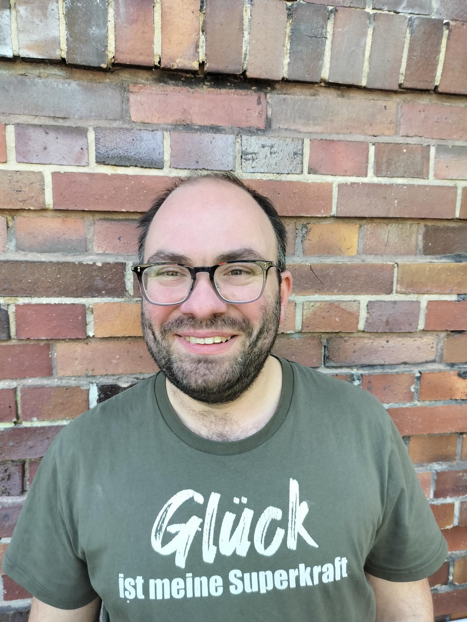 Richard trägt eine Brille mit schwarzem Rand und lächelt in die Kamera. Er hat kurze schwarze Haare. Auf seinem T-Shirt steht: Glück ist meine Superkraft.