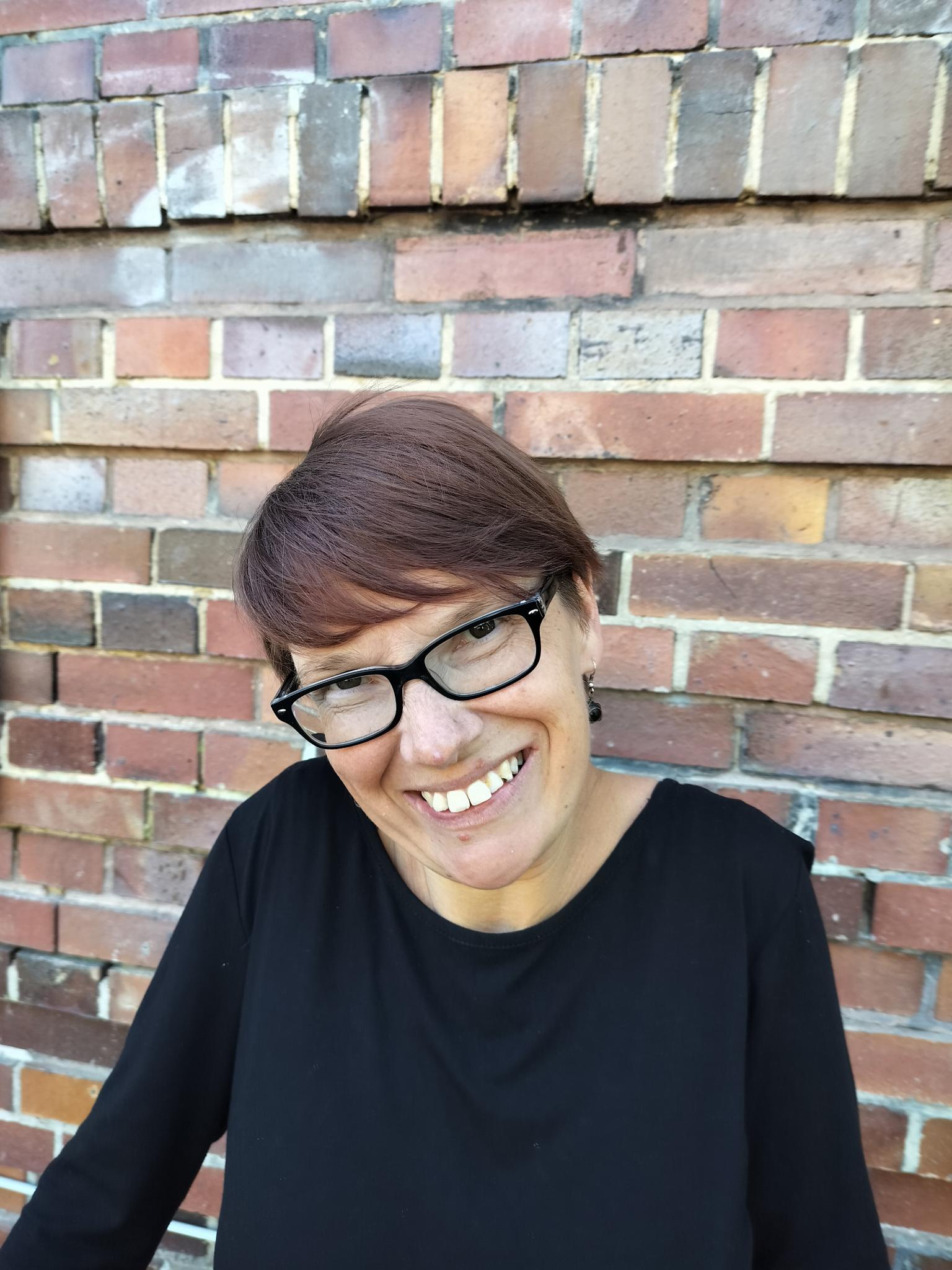 Anke trägt kurze rote Haare und eine schwarze, eckige Brille. Sie grinst und zeigt ihre Zähne. Sie steht vor einer Wand aus Backsteinen.